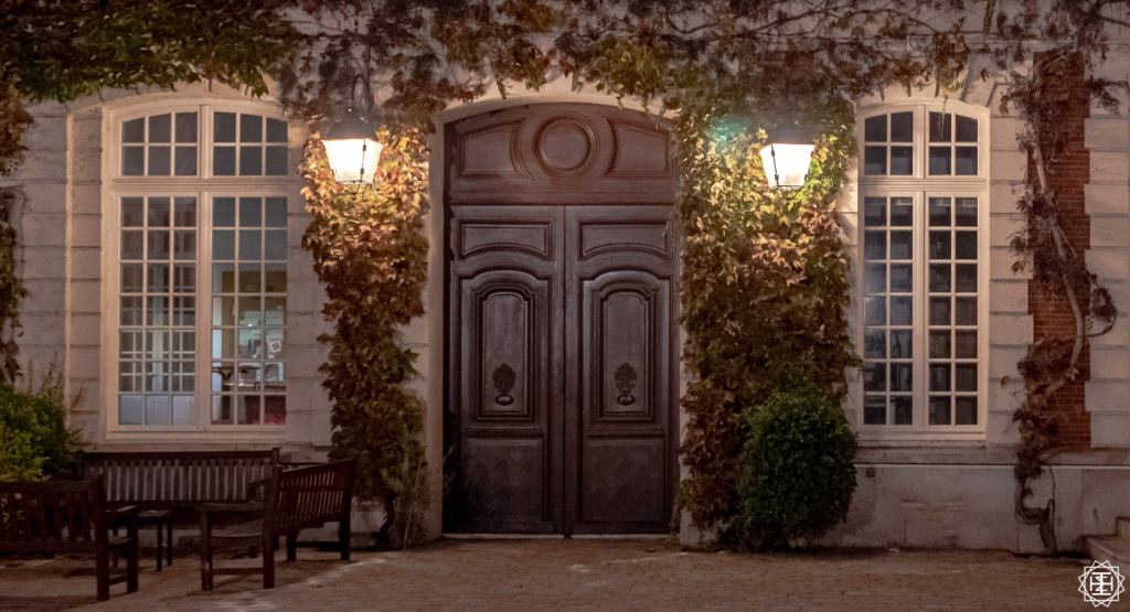 Closed doors at night with wall mounted lamps on each side. Lights inside of the building are off. The front is covered in ivy.